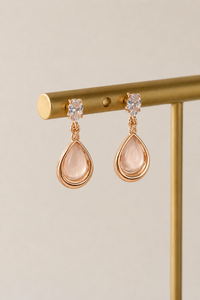 Rose gold teardrop earrings with clear gemstones on a gold stand against a beige background